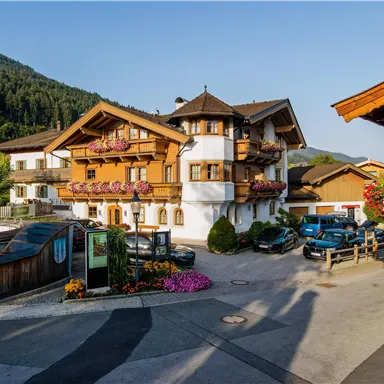 A picturesque chalet-like building with flowered balconies, surrounded by mountains. The road and parked cars add to the rural atmosphere.