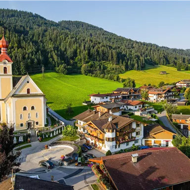 A picturesque mountain landscape with a church in the foreground and traditional houses. The green meadows and the wooded hills create a tranquil atmosphere.