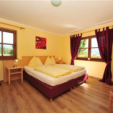 Ein gemütliches Schlafzimmer mit einem Doppelbett und gelben Wänden. Die Fenster bieten einen Blick auf die Natur, und Vorhänge in dunkelrot ergänzen die Einrichtung.