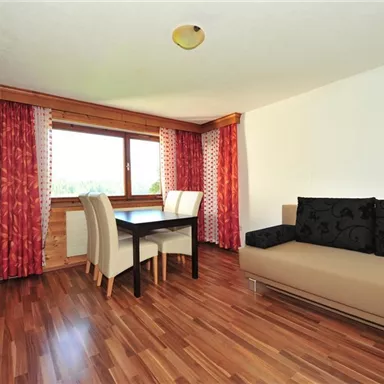 Ein helles Wohnzimmer mit Holzfußboden und modernen Möbeln. Es gibt einen Tisch mit Stühlen und eine Sofa-Ecke, die eine angenehme Atmosphäre schafft.
