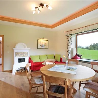 A bright living room with a cozy corner and a round wooden table. The large window view opens up to a green landscape.