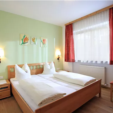 A cozy bedroom with a double bed and a light green wall color. The room has large windows with curtains and offers an inviting atmosphere.
