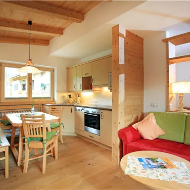 A bright, cozy apartment with a wooden beam ceiling. The furnishings include a kitchenette, a dining table, and a seating area with a red sofa.