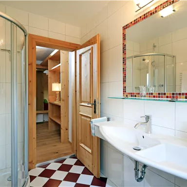 A modern bathroom with a shower, a sink, and wooden elements. The floor is designed with red and white tiles.