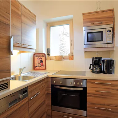 A modern kitchen with wooden cabinets and white countertops. It features appliances such as a stove, a microwave oven, and a coffee maker.