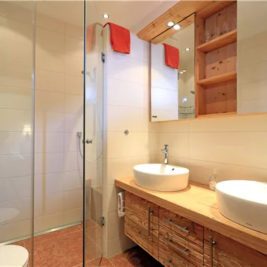A modern bathroom with a glass shower and two sinks. The furniture is made of wood and the walls are designed to be bright and cheerful.