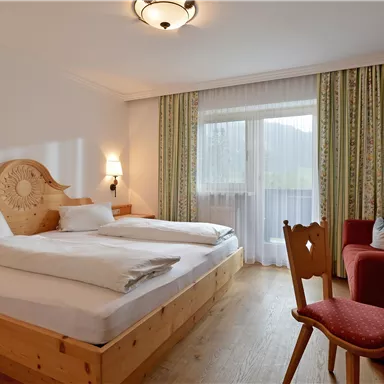 A cozy hotel room with wooden furniture and a double bed. There is a small table, a TV, and access to a balcony with a view outside.