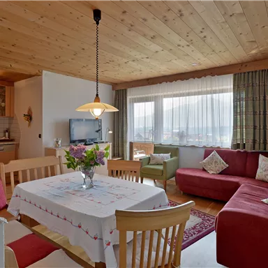 Ein gemütlicher Wohnbereich mit einem roten Sofa, einem Esstisch und großen Fenstern. Die Holzbalkendecke und die Blumen verleihen dem Raum eine warme Atmosphäre.