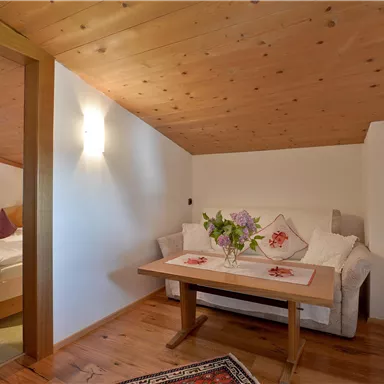 Ein gemütlicher Raum mit Holzdecken und einem Tisch in der Mitte. Auf dem Tisch stehen frische Blumen und nebenan befindet sich ein Schlafbereich.