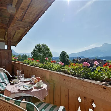 Ein gemütlicher Balkon mit Blick auf die Berge. Auf dem Tisch stehen Frühstücksplatten und Blumen blühen in den Kästen.
