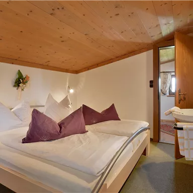 Ein gemütliches Schlafzimmer mit einem Doppelbett und lila Kissen. Die holzverkleidete Decke und die sanfte Beleuchtung schaffen eine warme Atmosphäre.