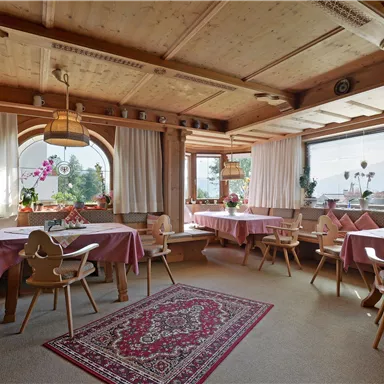 Ein gemütlicher Speiseraum mit Holzwänden und bunten Tischdecken. Große Fenster lassen viel Tageslicht herein und Pflanzen sorgen für eine einladende Atmosphäre.