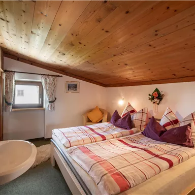 Ein gemütliches Schlafzimmer mit einem Dachboden aus Holz. Das Zimmer verfügt über ein Doppelbett mit karierten Bettdecken und einen angrenzenden Waschbereich.
