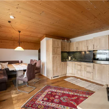 Eine gemütliche Küche mit Holzdetails und einem Esstisch für mehrere Personen. Der Wohnraum ist hell und freundlich mit einem Teppich und bequemen Sitzgelegenheiten.