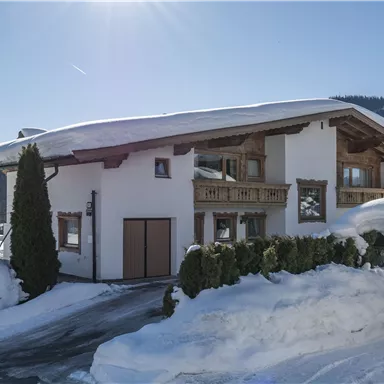 Ein schönes Haus im alpinen Stil, umgeben von schneebedeckten Hügeln. Die Sonne scheint unter einem klaren Himmel.