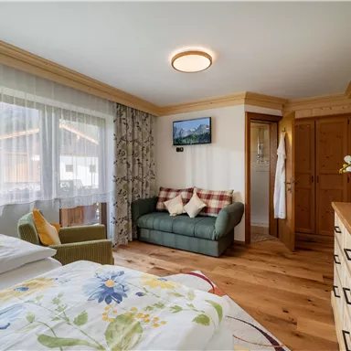 Ein helles Zimmer mit zwei Betten und einem gemütlichen Sofa. Große Fenster bieten Blick ins Freie und Holzdetails verleihen dem Raum eine warme Atmosphäre.