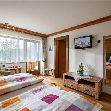 Ein gemütliches Schlafzimmer mit zwei Betten und bunten Bettbezügen. Der Raum hat große Fenster, einen Fernseher und eine moderne Holzkommode.