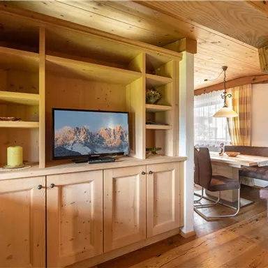 Ein gemütlicher Innenraum mit Holzregalen und einem Fernseher. Im Hintergrund ist ein Essbereich mit Stühlen und einem Tisch zu sehen.