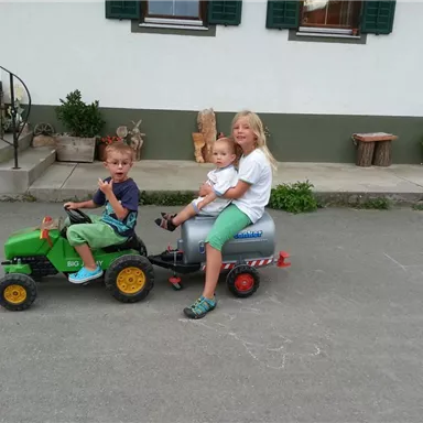 Drei Kinder fahren auf einem Spielzeugtraktor. Ein Junge fährt, while ein Mädchen und ein Kleinkind im Anhänger sitzen.
