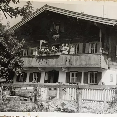 Ein traditionelles altes Haus mit Balkon und blühenden Pflanzen. Vor dem Haus stehen einige Personen und es ist von Bäumen umgeben.