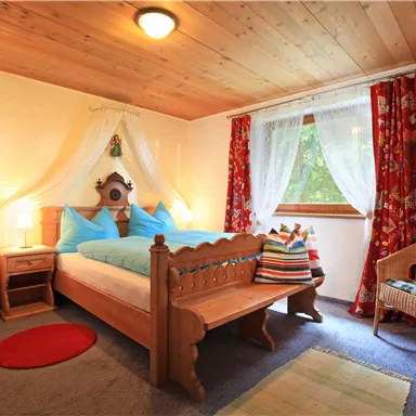 A cozy bedroom with a large bed and blue cushions. The room has wooden ceilings, colorful curtains, and a wicker chair.