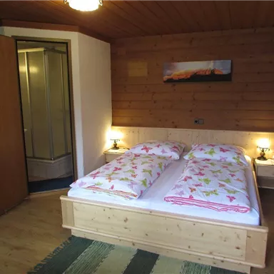 Ein gemütliches Schlafzimmer mit einem Doppelbett und floralem Bettzeug. Im Hintergrund ist ein Badezimmer mit Dusche vorhanden.