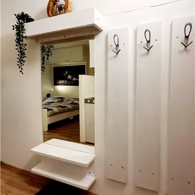 Ein moderner Eingangsbereich mit einem weißen Spiegel und Garderobenhaken. Im Hintergrund ist ein Schlafzimmer mit einem Doppelbett sichtbar.