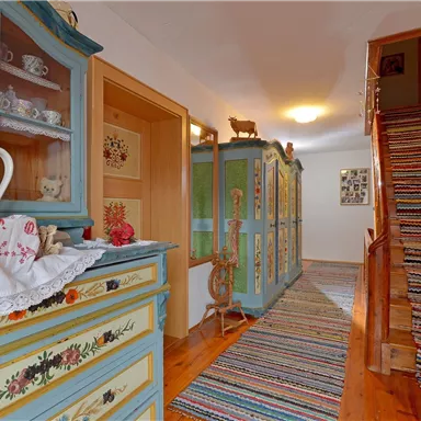 Ein gemütlicher Flur mit bunten Teppichen und hellen Wänden. Auf der linken Seite stehen dekorative Möbelstücke und eine Treppe führt nach oben.