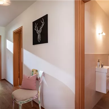 A bright hallway with a wooden door and a chair. Next to the chair is a small bathroom with modern decor.