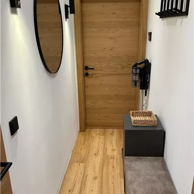 A narrow hallway with a wooden floor and a light wall color. There is a mirror, an entrance door, and a small seating area with a side table.