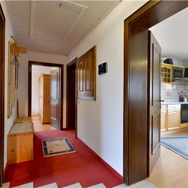A cozy hallway with wooden doors and a red carpet. In the background, a kitchenette is visible.