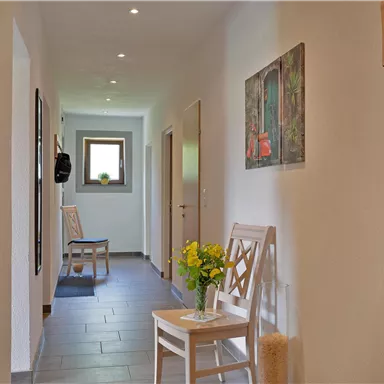 A bright hallway with white walls and a gray floor. On the left side, there are seating areas and a table with a flower vase.
