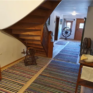 Ein einladender Flur mit einer Holztreppe und einem bunten Teppich. Auf der linken Seite steht ein antikes Möbelstück und eine Spinnmaschine.