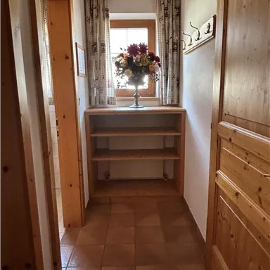 Ein heller Flur mit Holzwänden und einem Regal. Auf dem Regal steht eine Blumenvase, und durch das Fenster fällt Tageslicht.