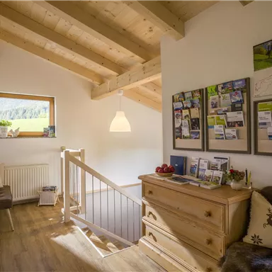 Ein heller Flur mit Holzdecke und Fenster, das viel Tageslicht hereinlässt. An der Wand hängen Informationskarten und ein Sideboard aus Holz steht im Raum.