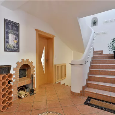 A bright hallway with a staircase and a plant in a pot. A decorative picture hangs on the wall, and a terracotta fireplace stands in the corner.