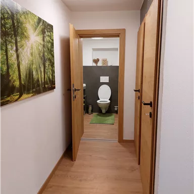 Ein heller Flur mit Holzfußboden und zwei offenen Türen. Im Hintergrund ist ein Badezimmer mit einer Toilette und grüner Badematte sichtbar.