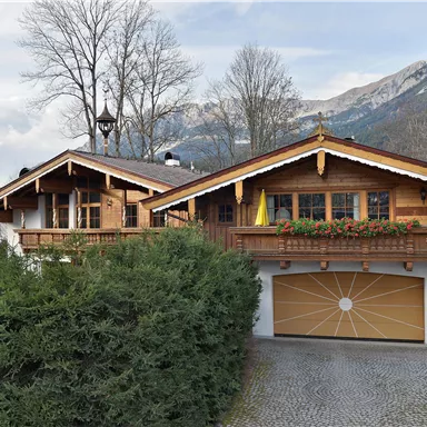 Ein charmantes Holzhaus im alpenländischen Stil, umgeben von Bäumen. Im Hintergrund sind majestätische Berge und ein bewölkter Himmel zu sehen.