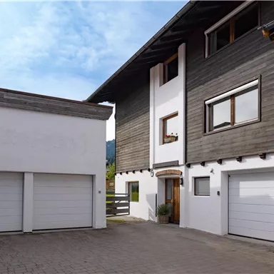 Ein modernes Haus mit einer Kombination aus Holz- und Putzfassade. Die Einfahrt hat zwei Garagentore und ist von einem klaren Himmel umgeben.