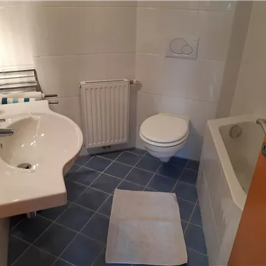 Ein modernes Badezimmer mit Waschbecken, Toilette und Badewanne. Der helle Raum hat graue Fliesen und eine orangefarbene Tür.