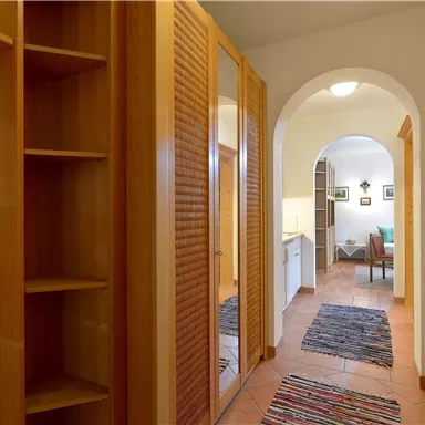 A bright corridor with a wooden cabinet and a carpet. In the background, another room is visible with a chair and picture frames.