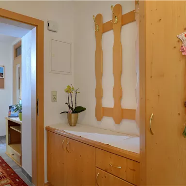 Ein helles Zimmer mit Holzschränken und einer Pflanze auf einem Tisch. Im Hintergrund ist ein weiterer Raum mit einem Stuhl und einer Pinnwand zu sehen.