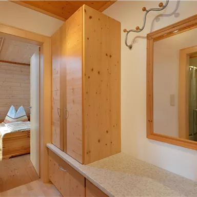 A bright hallway with wooden cabinets and a mirror. A cozy bedroom is visible in the background.