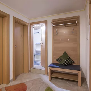 Ein heller Eingangsbereich mit Holzrahmen und einer Garderobe. Auf der Bank liegt ein grünes Kissen, und eine Tür führt zu einem weiteren Raum.
