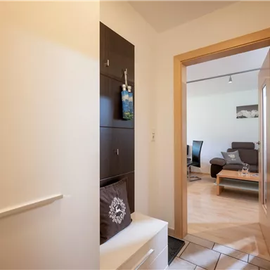A bright hallway with a shoe cabinet and an open door to the living room. In the background, a sofa and a table are visible.