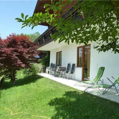 A cozy outdoor area of a house overlooking a well-maintained garden. Relaxed loungers and a red tree create an inviting atmosphere.