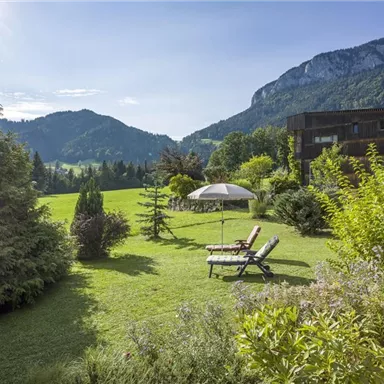 Eine ruhige Gartenlandschaft mit Sonnenliegen und einem Sonnenschirm. Im Hintergrund sind Berge und Bäume zu sehen, die eine malerische Kulisse bilden.