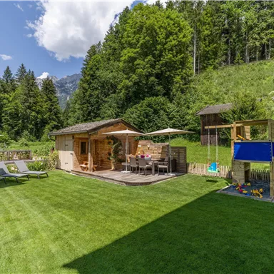 A beautiful garden with green grass and a wooden house. There are lounge chairs, a dining area, and a playground in the background.