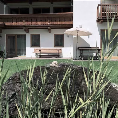 A tranquil garden with a stone fountain and lush greenery. In the background, there is a modern building with balconies and a sunshade.
