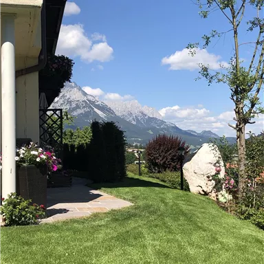 Eine malerische Aussicht auf Berge unter einem klaren blauen Himmel. Im Vordergrund befindet sich ein gepflegter Garten mit Blumen und Grünflächen.